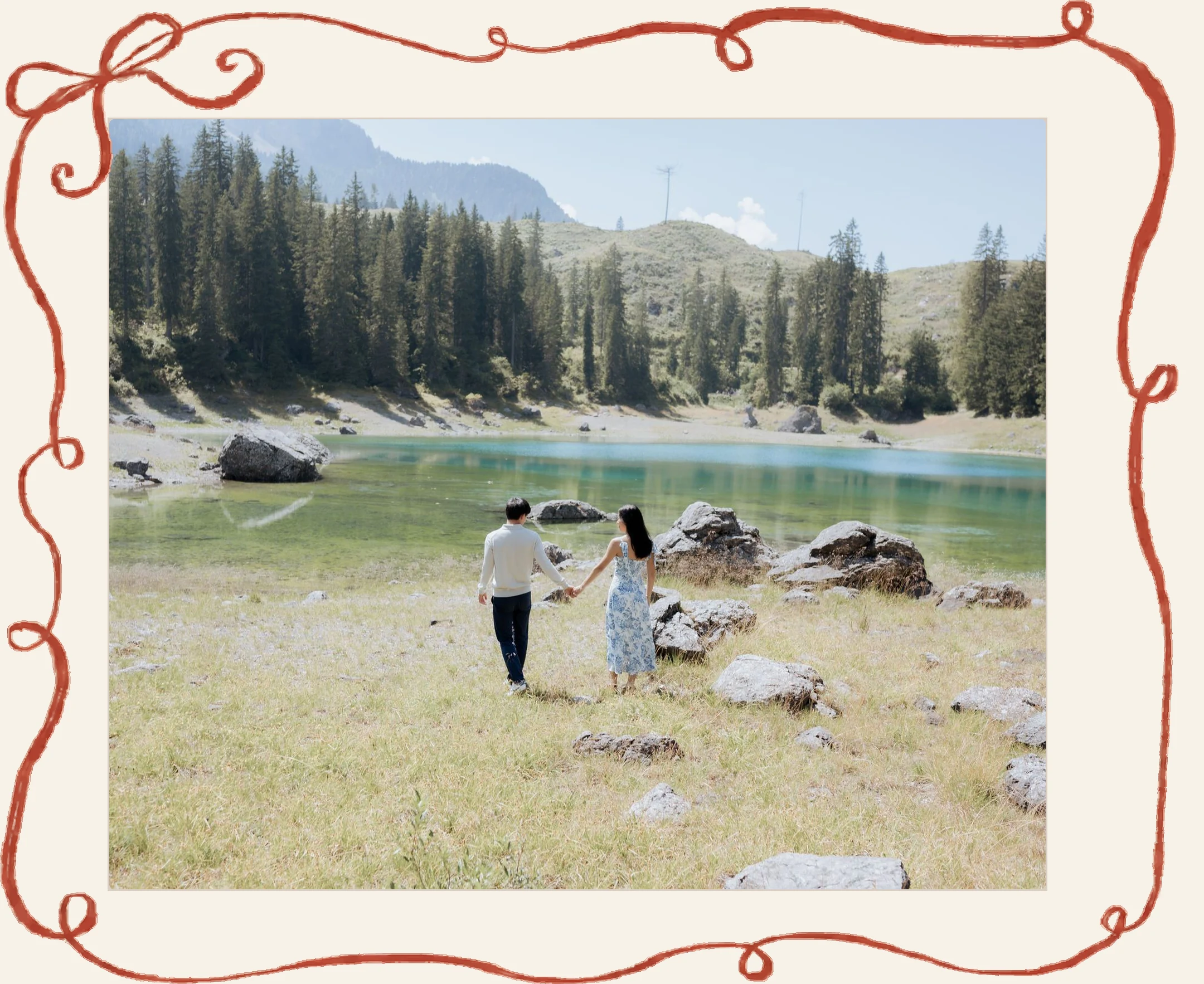Lia and Tony walking hand in hand along the shore of a turquoise mountain lake, mounted on cream paper inside YJ’s looping red ribbon frame.